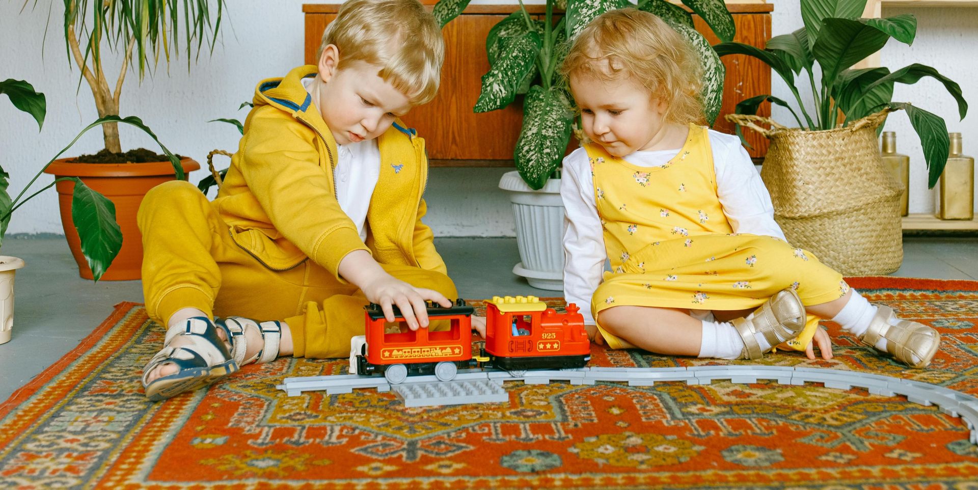 Two children enjoy playing with a toy train on a colorful rug indoors surrounded by plants.